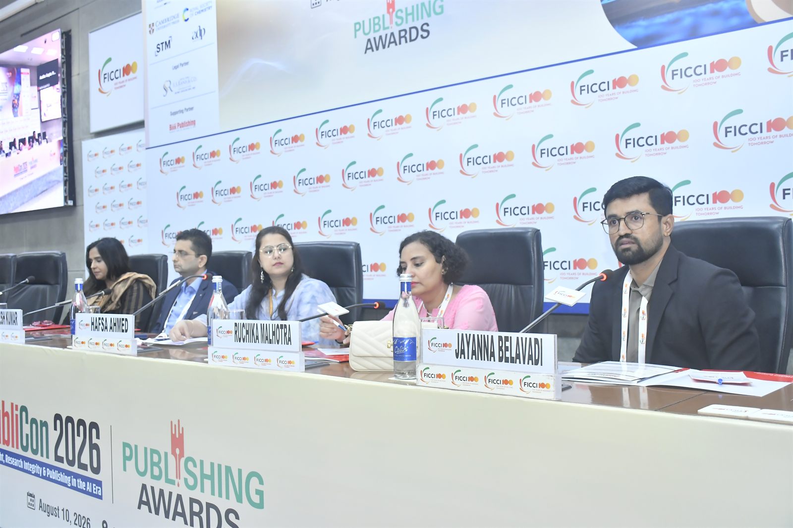 The PubliCon 2026 panel seated at the dais, with Jayanna Belavadi of DrillBit at the far end